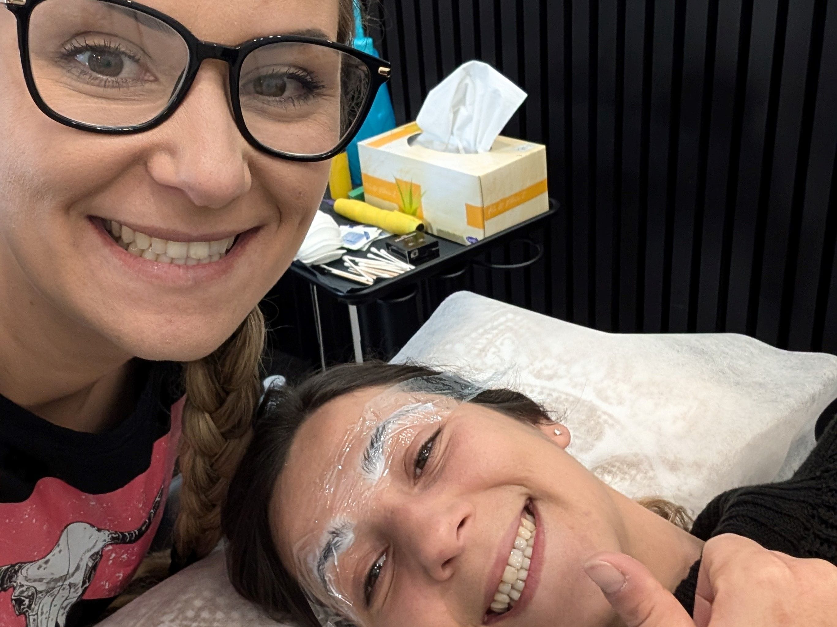 The owner and a woman smiling and giving a thumbs up while wearing numbing cream on her eyebrows before she receives her permanent eyebrow makeup, highlighting the use of top-quality products for customer safety.